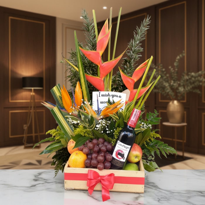 Arreglo floral Platonia con heliconias, aves del paraíso, uvas, manzanas, duraznos y botella de vino tinto, sobre mármol en u
