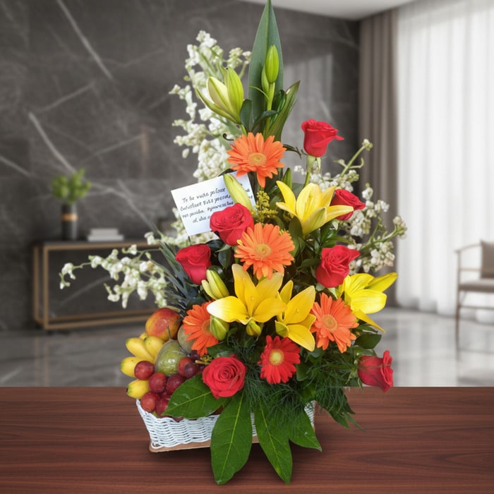 Arreglo floral con rosas rojas, lirios amarillos, gerberas naranjas y frutas frescas en una cesta blanca, sobre una mesa de m