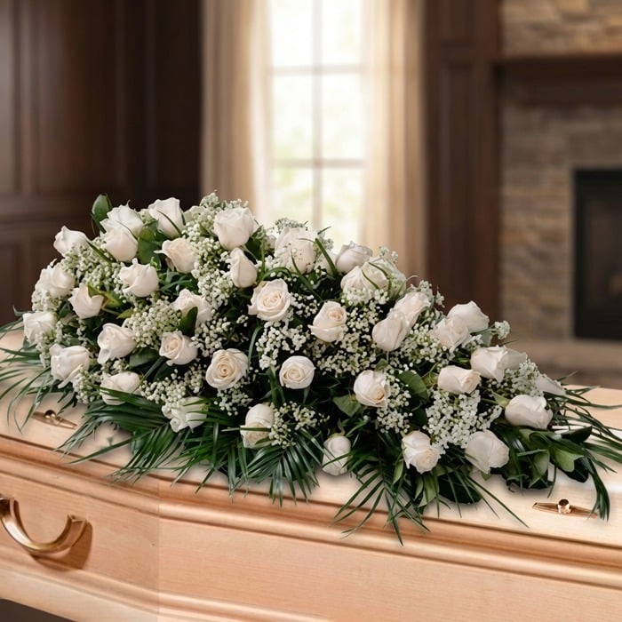 Arreglo fúnebre cubre caja con rosas blancas y follaje verde, sobre un ataúd de madera clara en una sala de velación premium