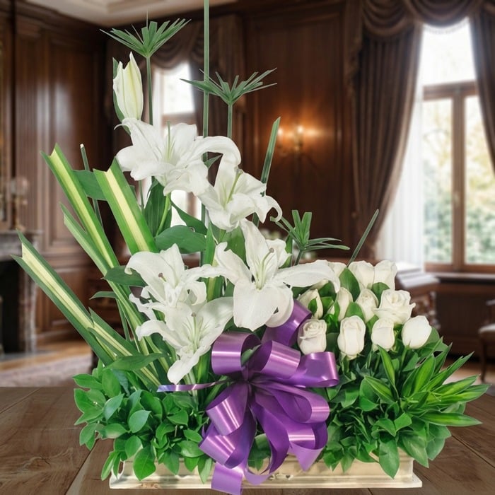 Arreglo fúnebre Pureza con lirios y rosas blancas, lazo morado, en sala de velación premium con luz natural y bokeh.