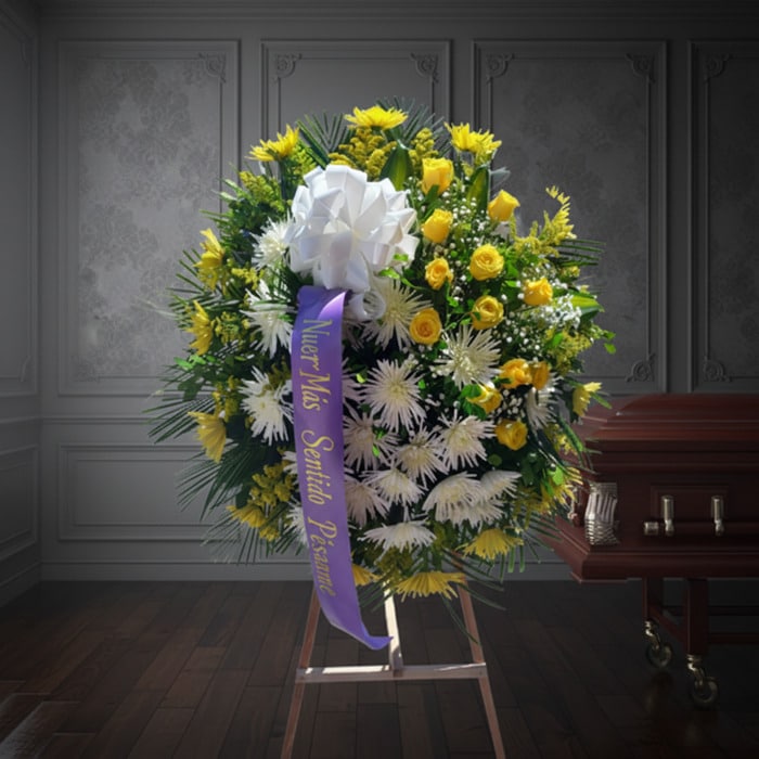 Corona fúnebre 'Estamos Contigo' con flores amarillas y blancas, cinta morada con mensaje de condolencia, junto a un ataúd en